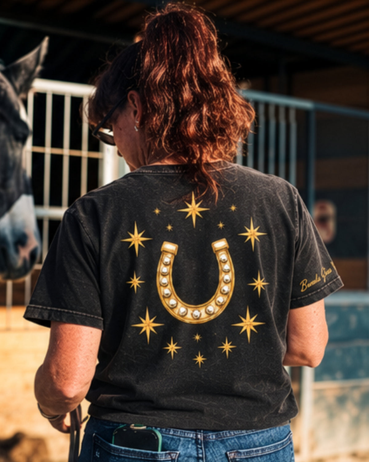 Icon Tee in black, back: a large gold horseshoe ringed with stars. The wearer stands at a stall beside a grey horse.