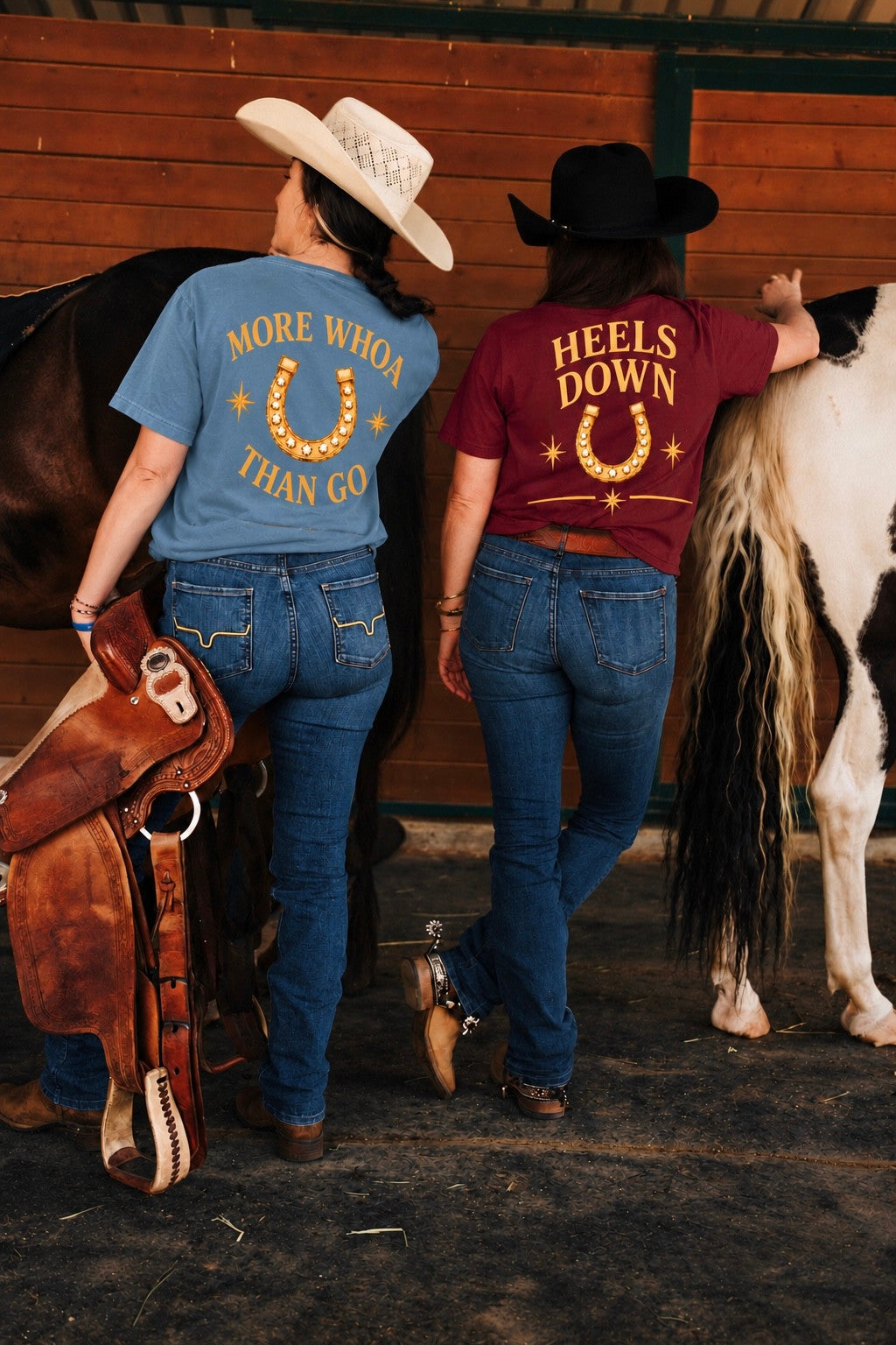 Two women from behind at a barn wall, in the More Whoa Than Go Tee in vintage denim and the Heels Down Tee in burgundy, a paint horse beside them.