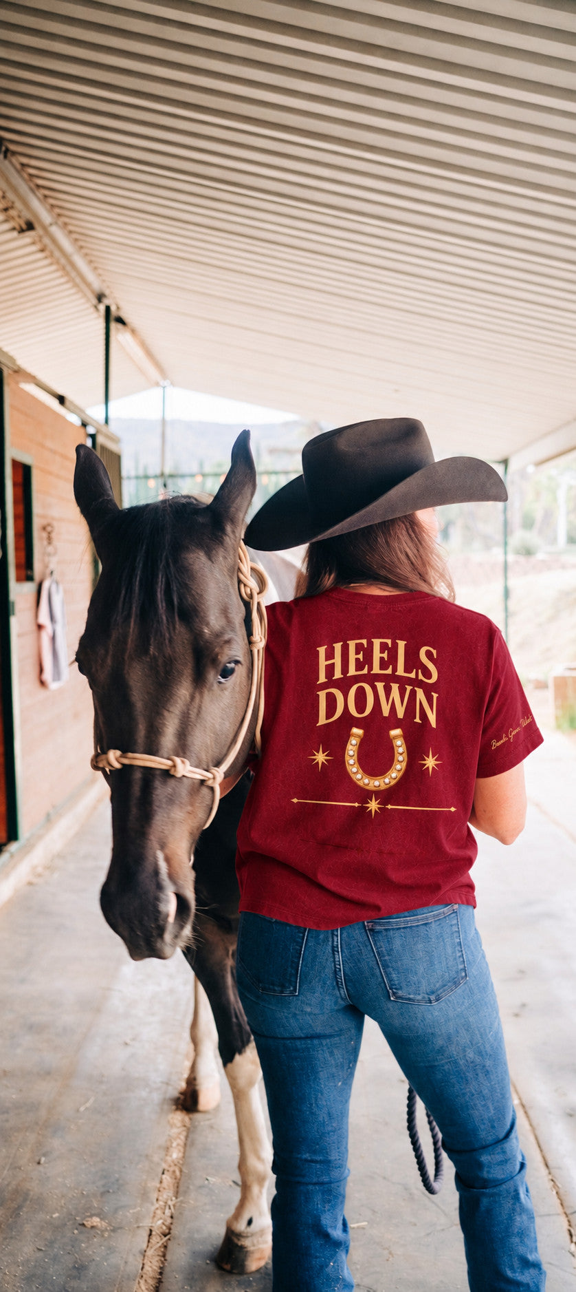 Heels Down Tee in burgundy, back: the words Heels Down above a gold horseshoe. The wearer walks beside a horse.