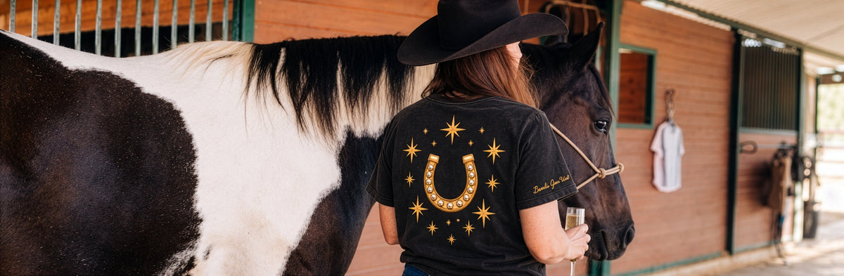 A rider in the black Icon Tee between a paint horse and a dark horse in a barn aisle.