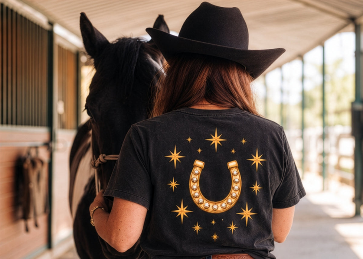 A rider in a black hat and the black Icon Tee beside a dark horse in a barn aisle.