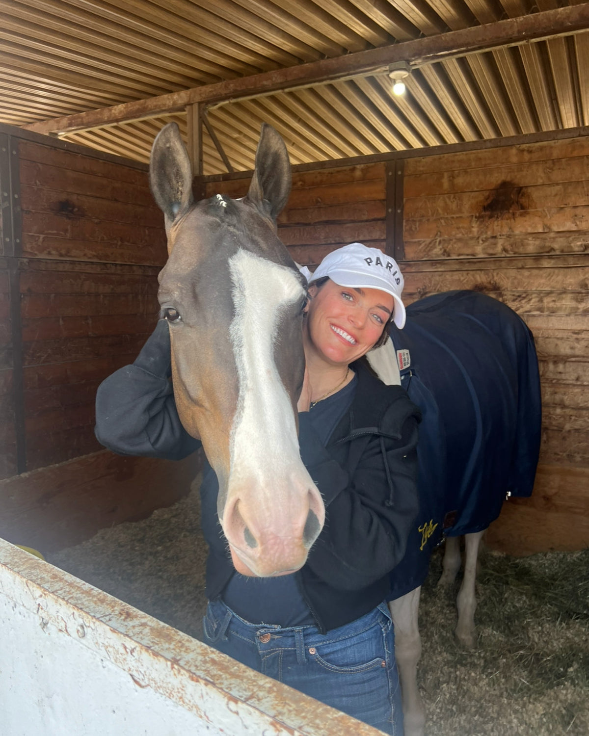 Kelly leaning her head against Bambi, a paint mare with a wide white blaze, in the mare's stall.